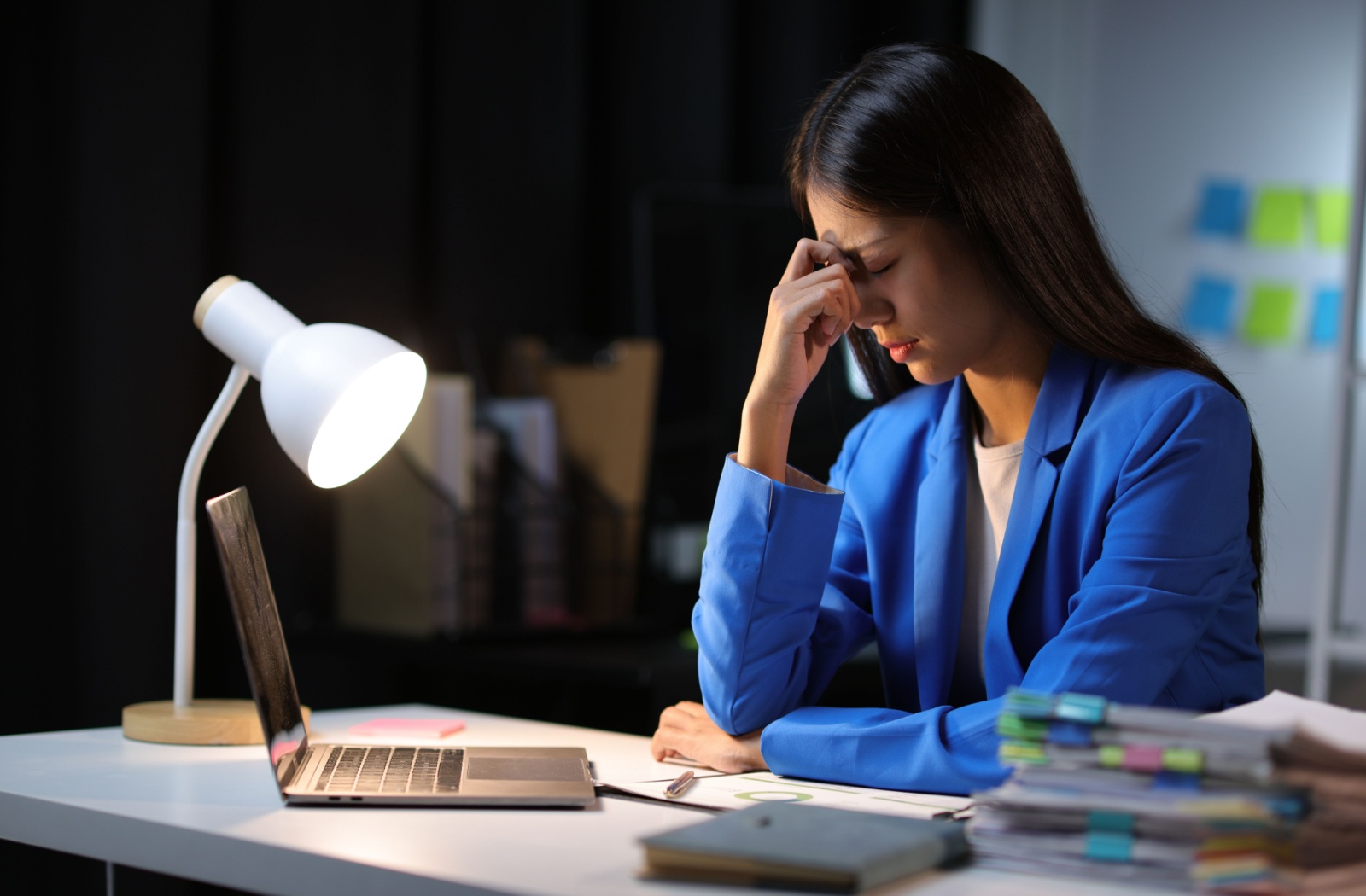 a patient wearing a blue blazer holds their forehead with their eyes closed to soothe symptoms of dry eyes and headache while working in front of their laptop.