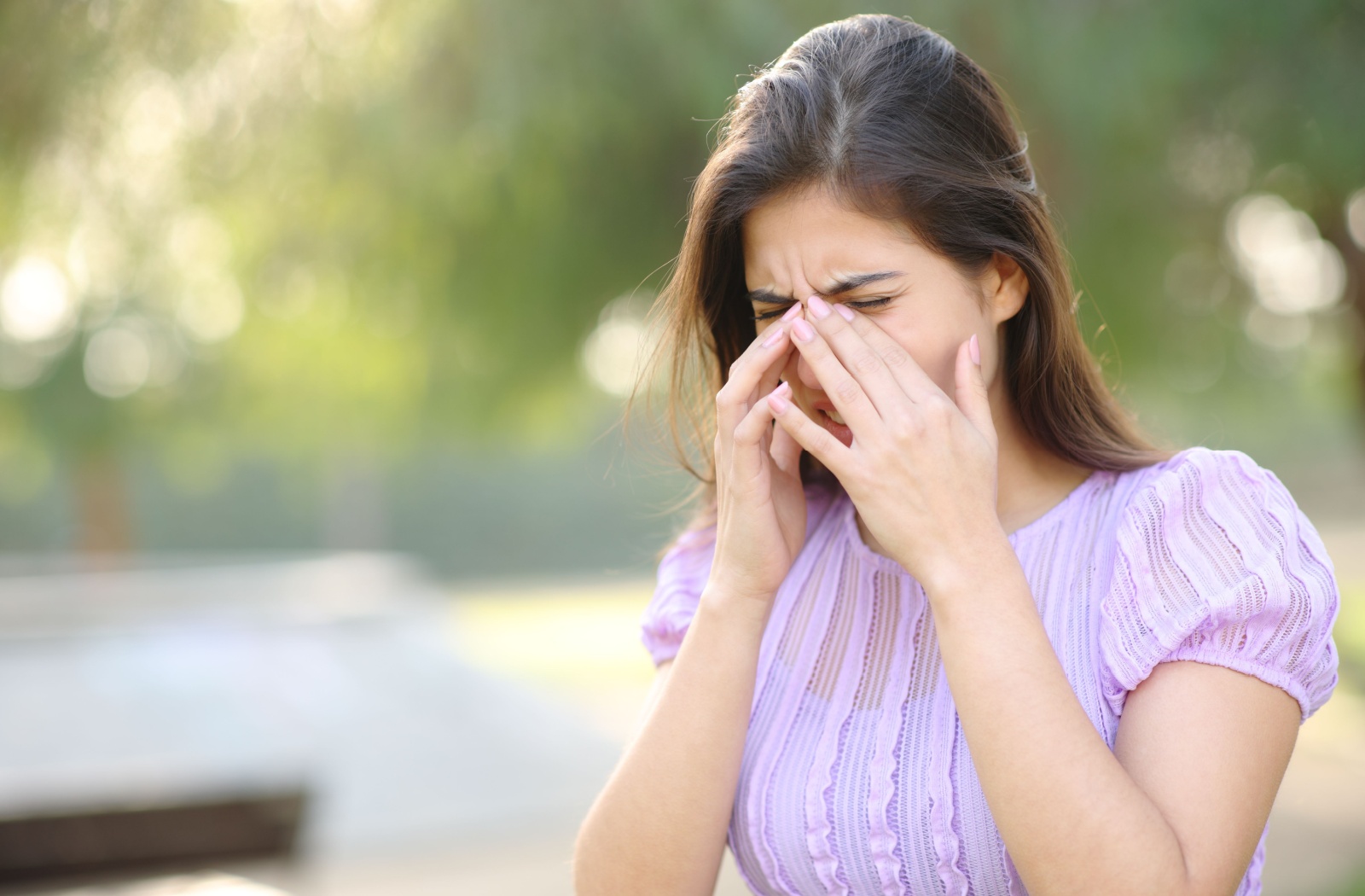 A woman outdoors winces in pain and rubs their eyes, either due to allergies or pink eye.