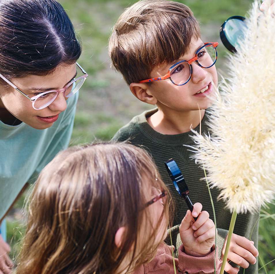 Three smiling children wearing eyeglasses looking at plant outdoors showing myopia control eyewear for active kids in natural setting.
