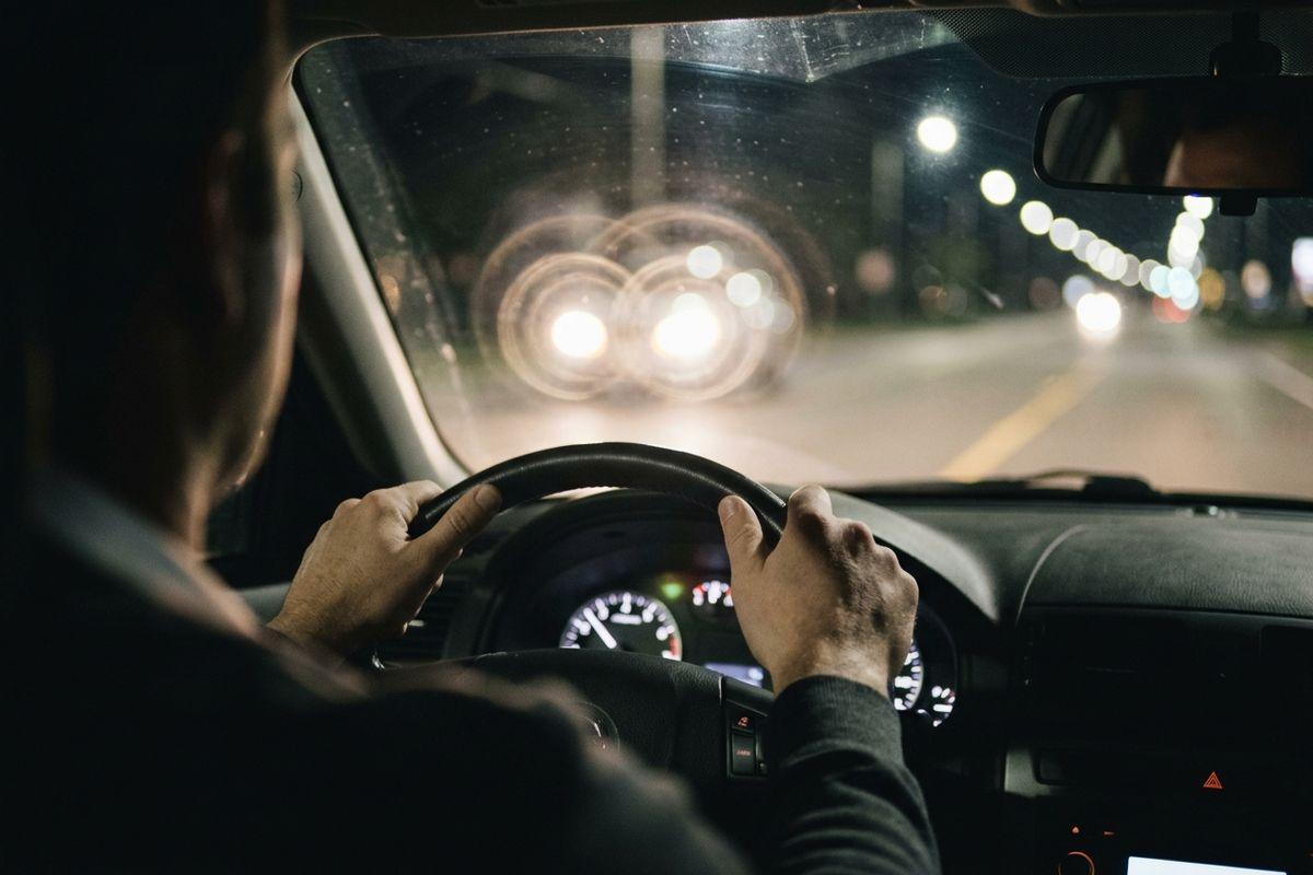 Adult gripping a steering wheel while driving at night, with oncoming headlights creating bright glare through the windshield.